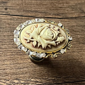 Può includere: Pomello per armadio ovale in stile antico con un design a cammeo di rosa. Il cammeo è color crema, su uno sfondo beige, ed è incorniciato da un bordo dorato. Strass trasparenti circondano il cammeo.