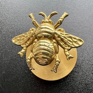 Può includere: Un battente per porta a forma di ape dorata. L'ape ha ali, antenne e segmenti del corpo dettagliati. È montato su una base rotonda dorata. Il battente è in metallo.