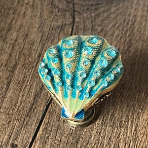 May include: A decorative seashell-shaped trinket box. The box is primarily teal and gold, with a fan-like design and small, sparkling blue gemstones. The box is set against a wooden background.