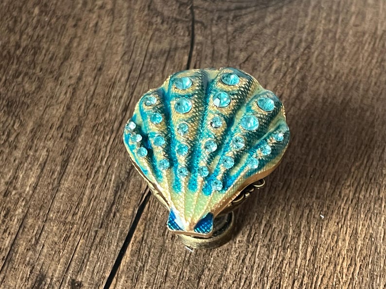 May include: A decorative seashell-shaped trinket box. The box is primarily teal and gold, with a fan-like design and small, sparkling blue gemstones. The box is set against a wooden background.