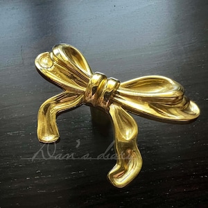 May include: Gold-toned metal bow-shaped cabinet knob or drawer pull. The bow is tied in a knot in the center.