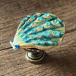 May include: Decorative seashell cabinet knob with a teal and gold finish, adorned with sparkling blue rhinestones. The knob has a bronze-coloured base and is designed for furniture or decor.