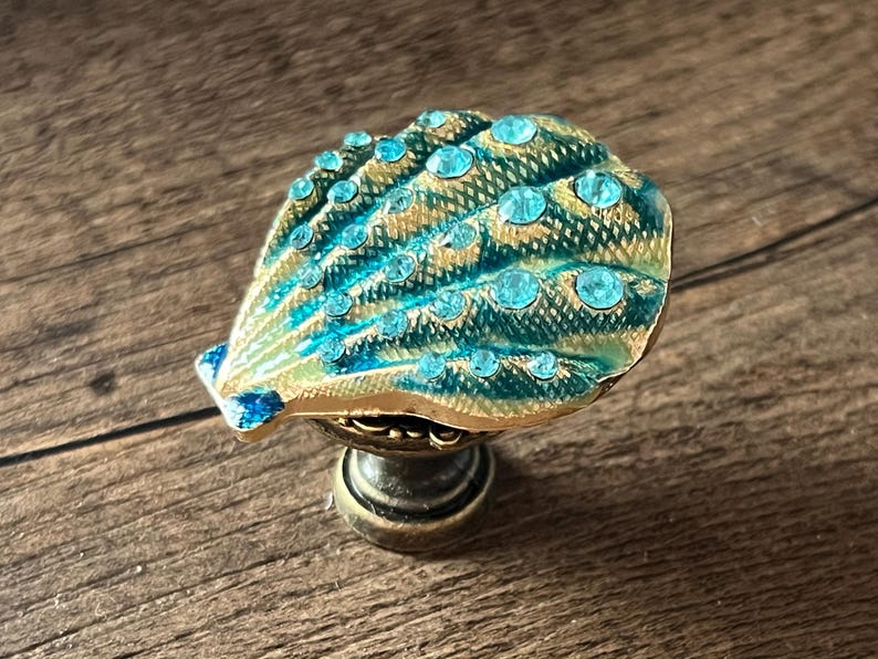 May include: Decorative seashell cabinet knob with a teal and gold finish, adorned with sparkling blue rhinestones. The knob has a bronze-coloured base and is designed for furniture or decor.