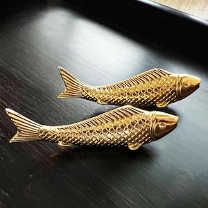 May include: Two gold-coloured fish-shaped decorative drawer pulls. The pulls are detailed with scales and have a shiny finish.