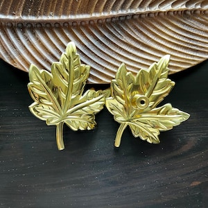 Könnte beinhalten: Zwei goldfarbene Dekorationsstücke in Form von Ahornblättern. Die Blätter sind detailliert und haben eine leicht strukturierte Oberfläche.