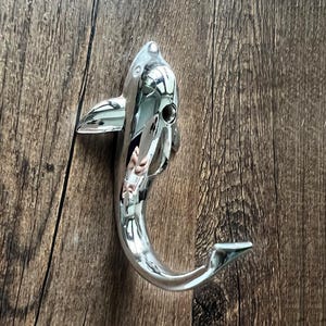 May include: A chrome-finished shark-shaped hook. The hook is mounted on a wooden surface. The shark has a curved body, a dorsal fin, and a tail fin. The hook is designed to hold items.