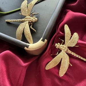 May include: Two gold-coloured dragonfly sculptures on a burgundy fabric background. One dragonfly is on a black tray with a gold trim.