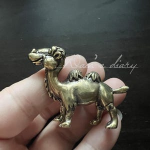 Puede incluir: Pequeña figura de camello de latón de estilo antiguo. El camello tiene un diseño detallado, incluyendo jorobas, patas y una cara sonriente. La figura se sostiene en una mano sobre un fondo oscuro. El texto "Nam's diary" está en el fondo.