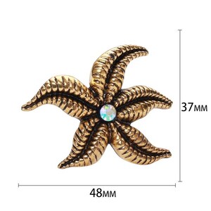 Op de afbeelding: Goudkleurige broche in de vorm van een zeester met een centrale iriserende edelsteen. De broche is 48 mm breed en 37 mm hoog. De zeester heeft gedetailleerde, getextureerde armen. Een decoratief accessoire.
