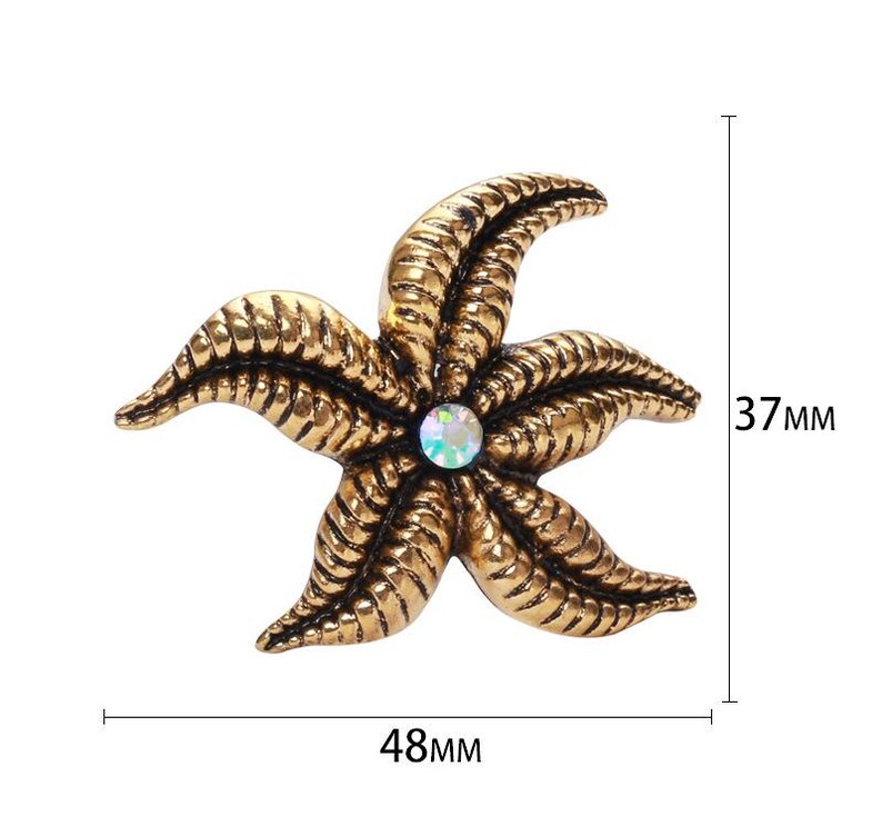 Op de afbeelding: Goudkleurige broche in de vorm van een zeester met een centrale iriserende edelsteen. De broche is 48 mm breed en 37 mm hoog. De zeester heeft gedetailleerde, getextureerde armen. Een decoratief accessoire.