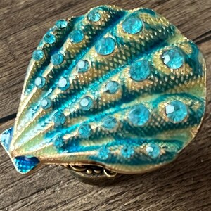 May include: A decorative seashell-shaped trinket box. The box is teal and gold with teal-coloured rhinestones. The box is on a wooden surface. The box is a decorative item.