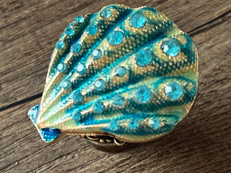 May include: A decorative seashell-shaped trinket box. The box is teal and gold with teal-coloured rhinestones. The box is on a wooden surface. The box is a decorative item.