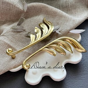 May include: Two gold leaf-shaped cabinet pulls with a decorative design. The pulls are on a white, textured surface.