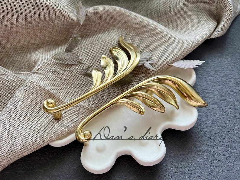 May include: Two gold leaf-shaped cabinet pulls with a decorative design. The pulls are on a white, textured surface.
