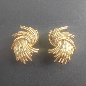 K&ouml;nnte beinhalten: Ein Paar goldfarbene Ohrstecker. Jeder Ohrring hat ein wirbelndes, f&auml;cherartiges Design mit einer strukturierten, gepunkteten Oberfl&auml;che. Die Ohrringe sind vor einem dunkelgrauen Hintergrund platziert.