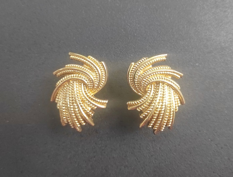 K&ouml;nnte beinhalten: Ein Paar goldfarbene Ohrstecker. Jeder Ohrring hat ein wirbelndes, f&auml;cherartiges Design mit einer strukturierten, gepunkteten Oberfl&auml;che. Die Ohrringe sind vor einem dunkelgrauen Hintergrund platziert.