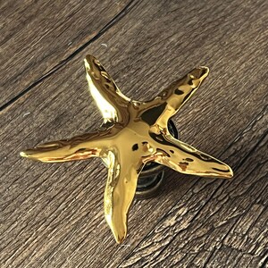 Puede incluir: Un tirador de armario dorado en forma de estrella de mar. El tirador tiene cinco puntas y una superficie texturizada. El herraje está montado sobre una base circular oscura. El fondo es una superficie de madera.