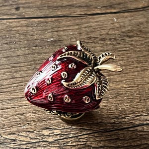 Puede incluir: Pomo decorativo en forma de fresa, rojo con detalles dorados. El pomo metálico presenta hojas y puntos dorados. Diseño vintage para muebles o decoración.