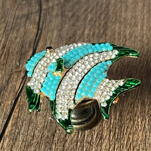 以下が含まれることがあります： ターコイズとクリアクリスタルアクセントが施された、魚の形をした装飾的なブローチ。ブローチは、グリーンのエナメルアウトラインとゴールドトーンのメタルベースが特徴です。魚のデザインは詳細で人目を引き、どんな服装にも輝きを加えます。