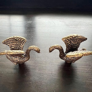 May include: A pair of bronze-coloured swan figurines with outstretched wings. The swans have detailed feather textures and curved necks. They are displayed on a dark wooden surface, showcasing their decorative design.