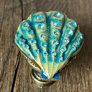 May include: A decorative seashell-shaped object in teal and gold. The shell is embellished with numerous clear, round rhinestones. The base is metallic gold, and the object is set against a wooden surface.