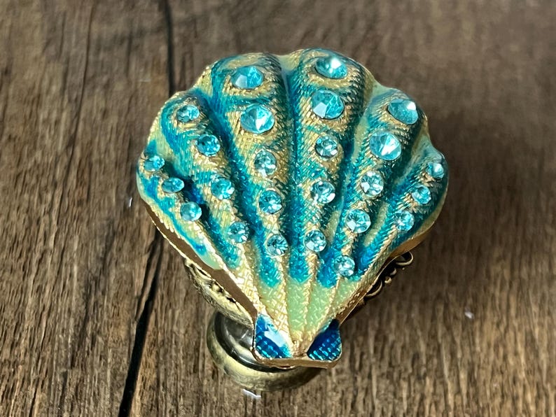 May include: A decorative seashell-shaped object in teal and gold. The shell is embellished with numerous clear, round rhinestones. The base is metallic gold, and the object is set against a wooden surface.