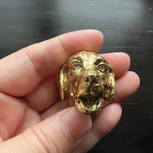 May include: A detailed, gold-coloured dog head sculpture with an open mouth and visible teeth. The sculpture is held in a hand, showcasing its intricate features and metallic sheen. The dog's expression appears alert and attentive.