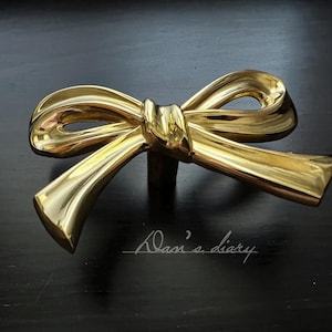 May include: A gold-colored bow-shaped cabinet pull. The hardware is made of metal and has a shiny finish. The bow is tied in the center with two loops on each side. The text "Dasi's diary" is written in cursive on the surface.