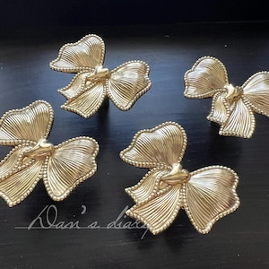May include: Four gold-colored bow-shaped cabinet pulls. Each pull features a detailed, ridged design and a small heart-shaped accent in the center. The pulls are arranged on a dark surface.