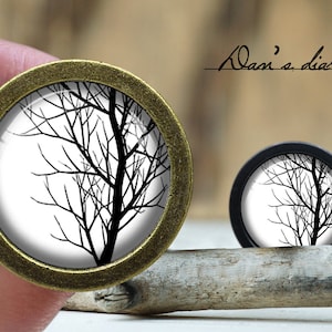May include: Two round cabinet knobs with a black and white tree branch design. One knob is held by a hand, and the other rests on a wooden stick. The knobs have a bronze and black finish. The text "Adam's diary" is written in cursive.