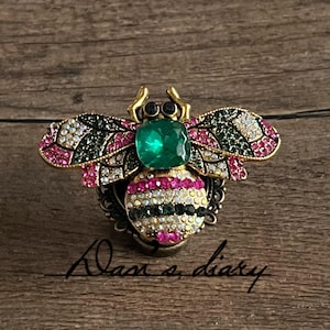 Può includere: Spilla a forma di ape gioiello con una grande pietra centrale verde smeraldo e ali decorate con cristalli rosa, verdi e bianchi. Il corpo dell'ape presenta strisce alternate di cristalli rosa, bianchi e neri. La spilla è su una base di metallo scuro.