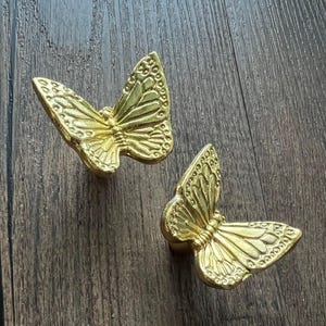 May include: Two gold butterfly cabinet knobs. The knobs are made of metal and have detailed wing patterns. The knobs are displayed on a dark wood surface.