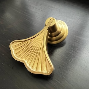 Puede incluir: Un llamador de puerta de metal dorado con forma de hoja de ginkgo. El llamador tiene un diseño detallado y texturizado y una base redonda.