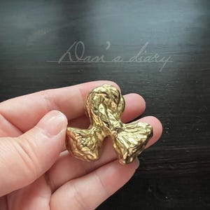 Könnte beinhalten: Eine goldfarbene Schleife aus Metall in Form eines Knotens. Die Schleife wird in einer Hand gehalten.