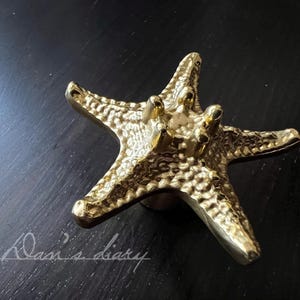 May include: A gold-coloured starfish cabinet knob. The knob has a textured surface and is mounted on a round base. The starfish has five points and is made of a shiny, metallic material. The background is a dark surface.