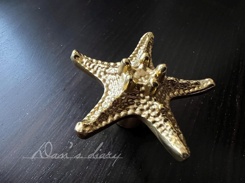 May include: A gold-coloured starfish cabinet knob. The knob has a textured surface and is mounted on a round base. The starfish has five points and is made of a shiny, metallic material. The background is a dark surface.