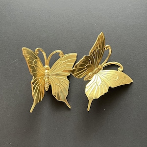 May include: Two gold-coloured metal butterfly wall decor pieces. The butterflies are facing different directions and have intricate details.