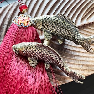 以下が含まれることがあります： 木製の表面に、詳細な鱗とひれを持つ2つのブロンズ色の鯉の置物があります。鮮やかな赤いタッセルと花のアクセントが付いた装飾的なチャームも存在します。鯉は泳いでいるかのように配置されています。