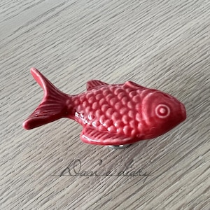 May include: A red ceramic fish figurine with a detailed scale pattern. The fish has a slightly upturned mouth and a large, round eye.