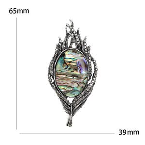 May include: An oval brooch with an abalone shell centre, set in an ornate silver-toned frame. The brooch is 6.5 cm long and 3.9 cm wide. The design features a detailed, textured pattern with small crystals, creating a vintage aesthetic. A statement piece.