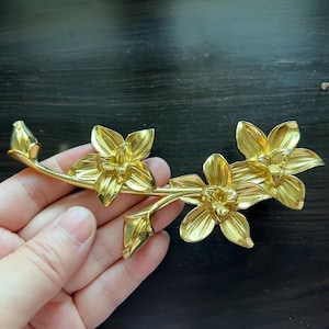 Pode incluir: Um broche de metal dourado em forma de ramo de flor com três flores. As flores têm um design detalhado e texturizado.
