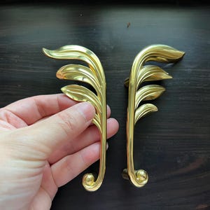 May include: Two gold-toned leaf-shaped cabinet pulls with a decorative scroll design at the bottom.