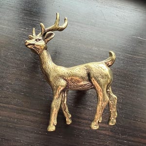 Op de afbeelding: Een messing beeldje van een hert, staand op vier poten met geweien. Het hert is gedetailleerd met een gladde, gepolijste afwerking. Het beeldje is ongeveer 15 cm hoog.