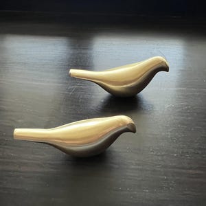 K&ouml;nnte beinhalten: Zwei goldene Vogelfiguren aus Metall auf einer dunklen Holzoberfl&auml;che. Die V&ouml;gel sind in die gleiche Richtung ausgerichtet und haben einen einfachen, modernen Stil.