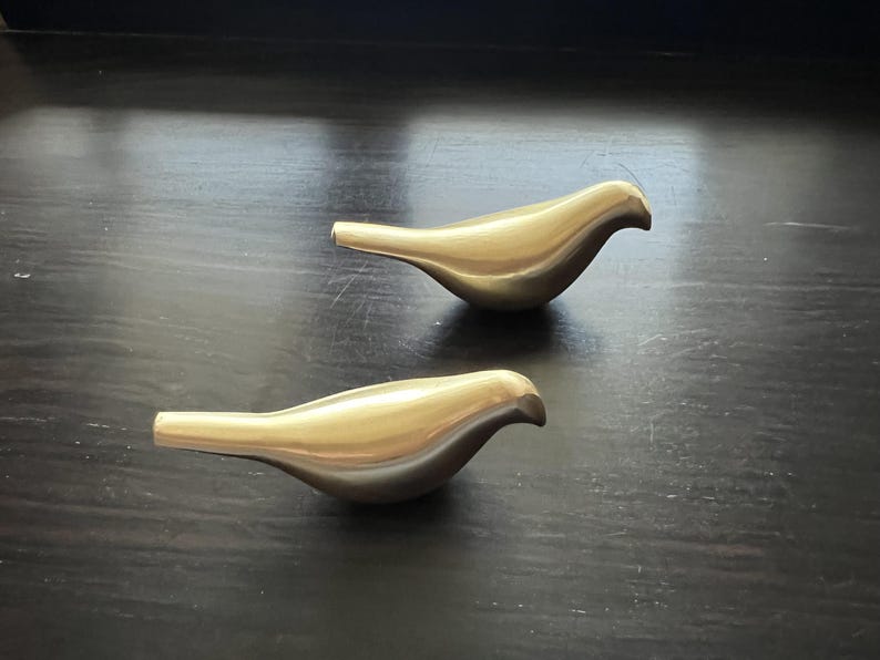 K&ouml;nnte beinhalten: Zwei goldene Vogelfiguren aus Metall auf einer dunklen Holzoberfl&auml;che. Die V&ouml;gel sind in die gleiche Richtung ausgerichtet und haben einen einfachen, modernen Stil.