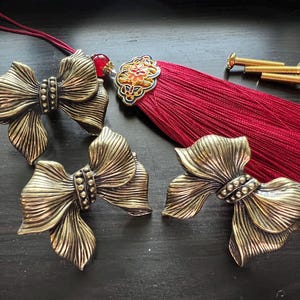 May include: Bronze-coloured bow-shaped drawer pulls and a red tassel with decorative accents. The drawer pulls have a detailed, textured design. Gold-coloured screws are also visible.