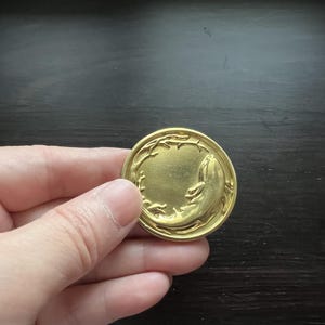 May include: A gold-coloured metal coin with a raised design of a fish jumping out of a circle of leaves.