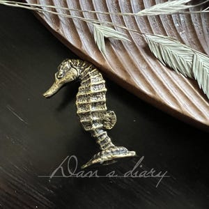 Pode incluir: Uma figura detalhada de cavalo-marinho em latão, em estilo antigo. O cavalo-marinho está posicionado sobre uma superfície escura, perto de uma bandeja de madeira e folhagem seca. O cavalo-marinho tem aproximadamente 7.6 cm de altura. A imagem tem uma estética vintage e quente.