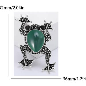 以下が含まれることがあります： 緑色の楕円形の中心石と黒いアクセントが付いた、銀色のカエルのブローチ。ブローチのサイズは長さ52mm、幅36mmです。カエルの体は、小さなテクスチャード加工されたドットで詳細に表現されています。
