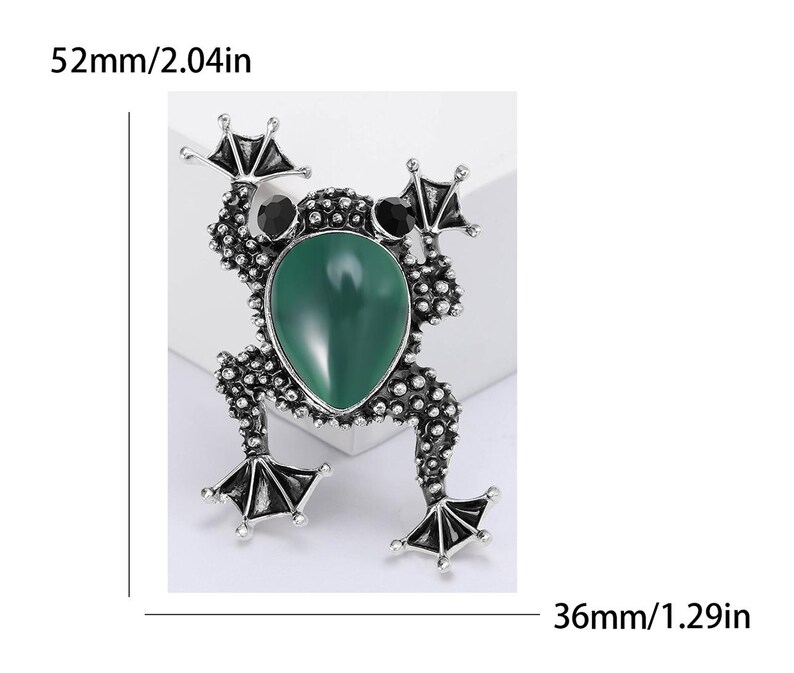 以下が含まれることがあります： 緑色の楕円形の中心石と黒いアクセントが付いた、銀色のカエルのブローチ。ブローチのサイズは長さ52mm、幅36mmです。カエルの体は、小さなテクスチャード加工されたドットで詳細に表現されています。